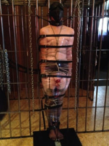 Dominatrix Domina Yuki has a male submissive tied up and bound in layered bondage while standing.