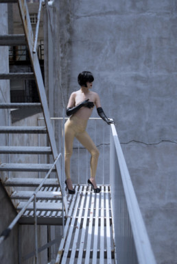Professional Dominatrix Domina Yuki stands on a fire escape elegantly dressed and covering her breasts with gloved hands.